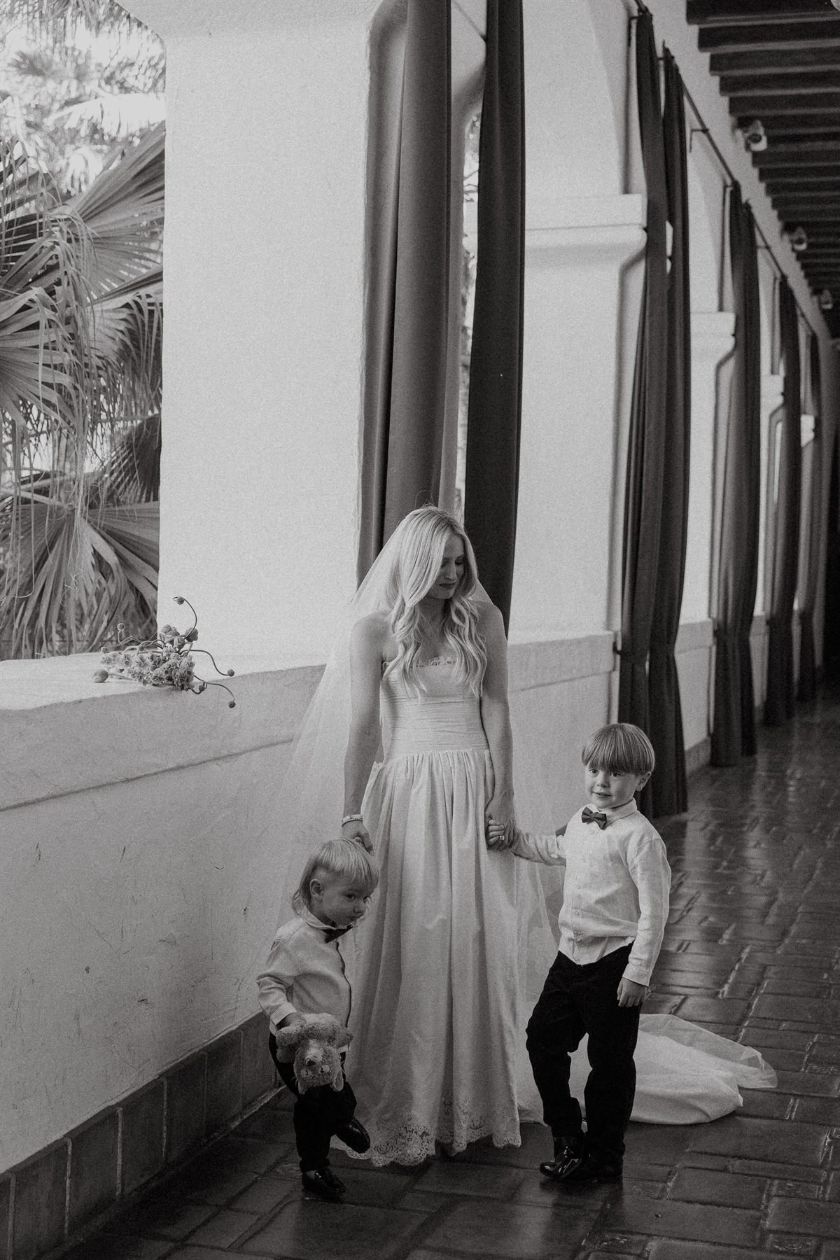 Black-and-white frame of Brittany Casta&ntilde;os walking the courthouse colonnade holding hands with her two sons in their suits.