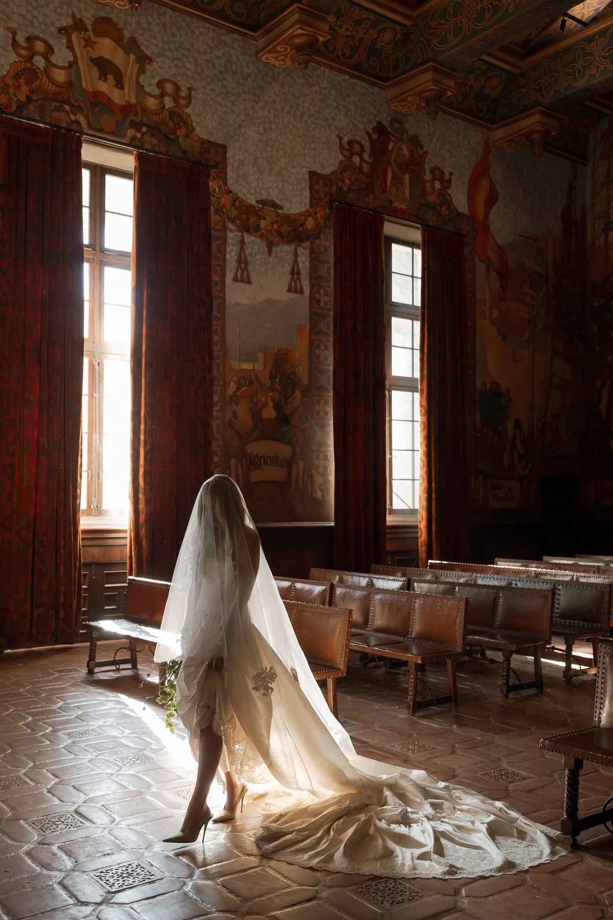 The ceremony look, the Montecito gown and Mural veil by Daughters of Simone, seen from behind as the bride walks through the Mural Room.