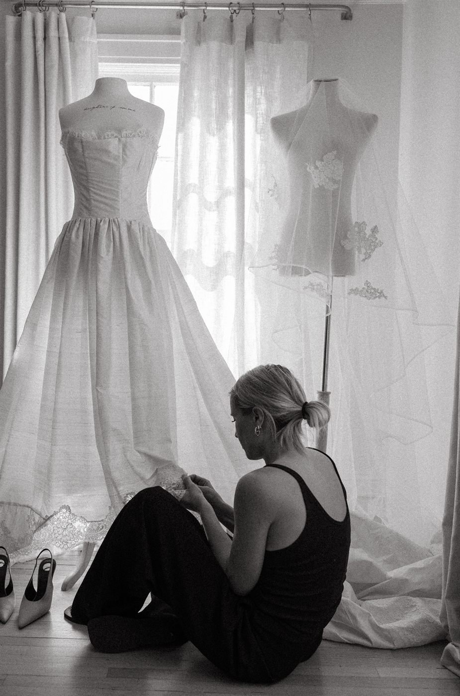 Black-and-white frame of the designer sitting on the floor in front of her own wedding dress on its stand.