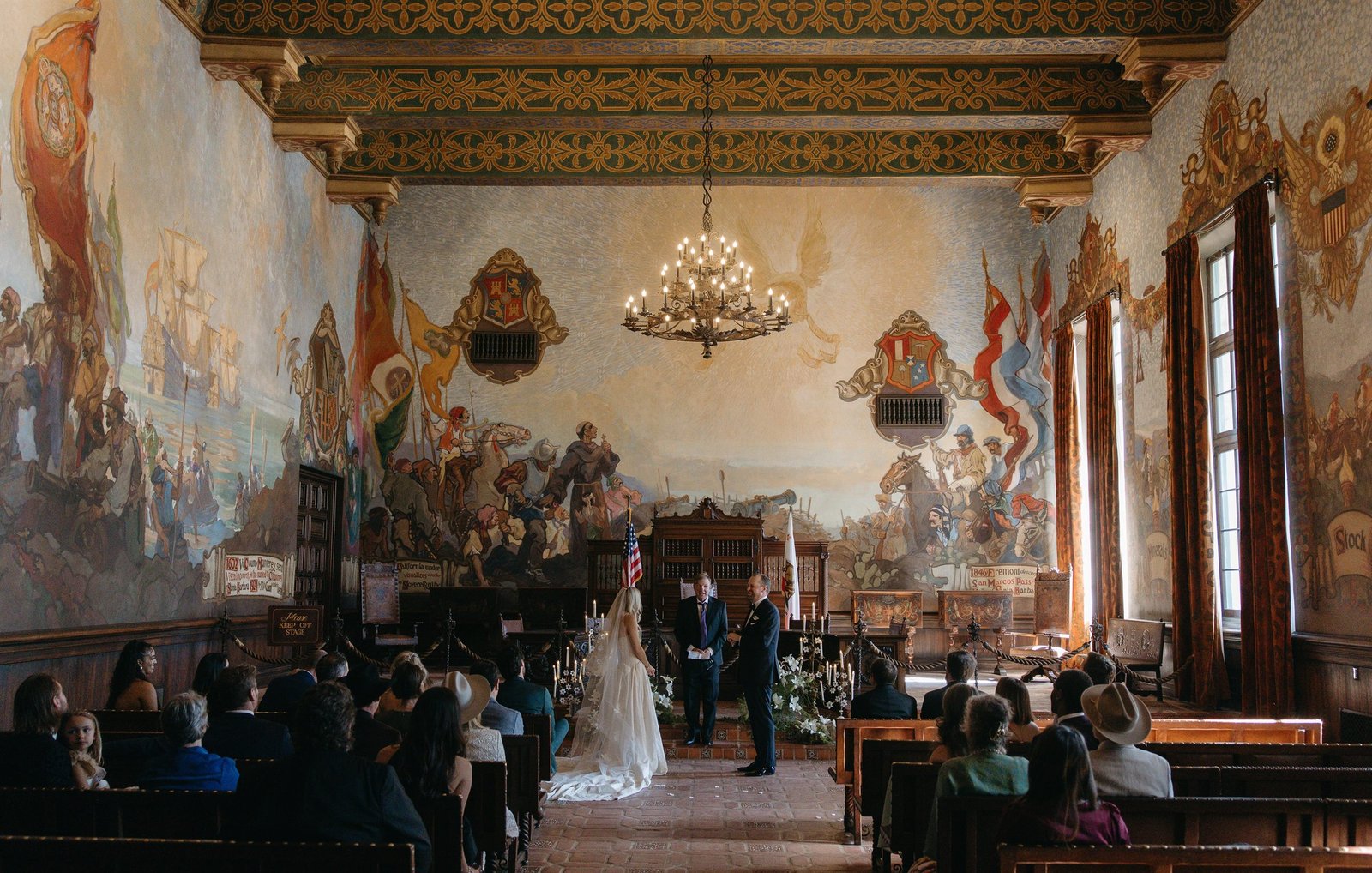 Wide view of the Mural Room at the Santa Barbara Courthouse during the ceremony, thirty guests seated beneath the painted ceiling and chandelier.